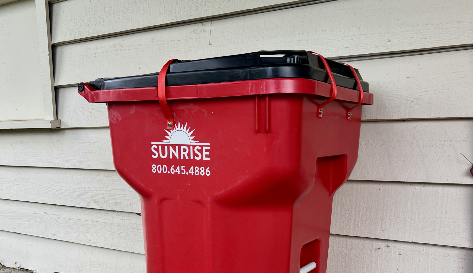 Red wheeled garbage bin with black lid against beige house siding; Sunrise logo and phone number on the side.] ,