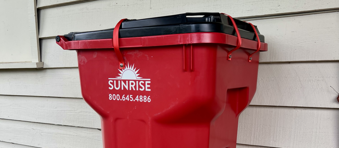 Red wheeled garbage bin with black lid against beige house siding; Sunrise logo and phone number on the side.] ,